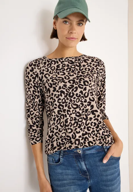 Frau mit grünem Cap, Leopardenprint-Shirt und blauer Jeans steht an der Wand.